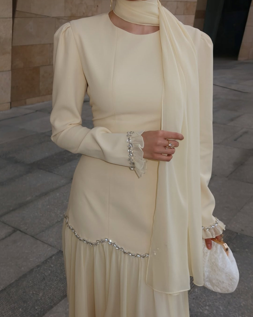 Woman wearing a cream-colored outfit with a scarf and jewelry on a stone pavement.