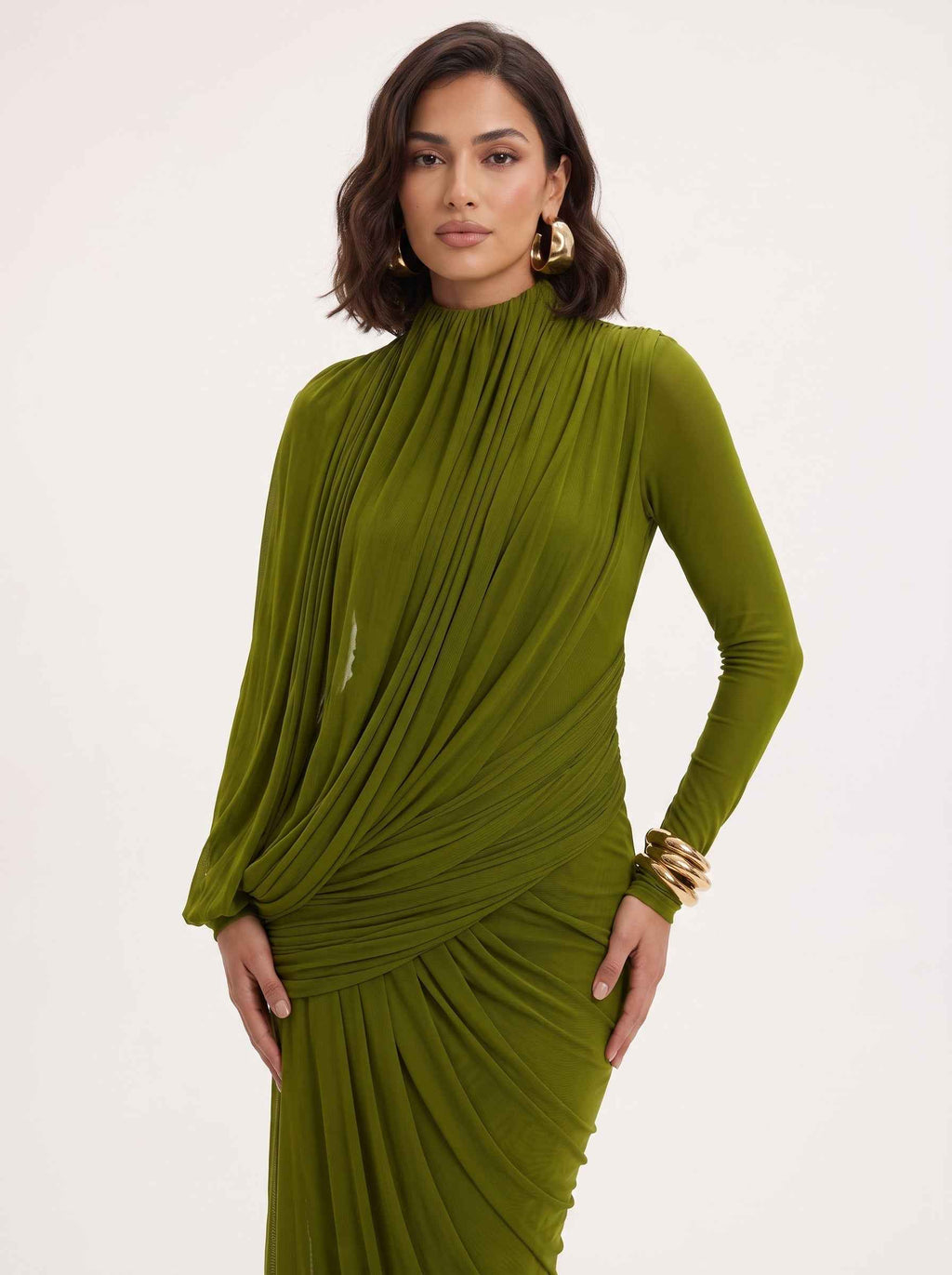 A woman modeling a draped olive green long-sleeve dress with a high mock neck and gathered pleated detailing, set against a plain light background.