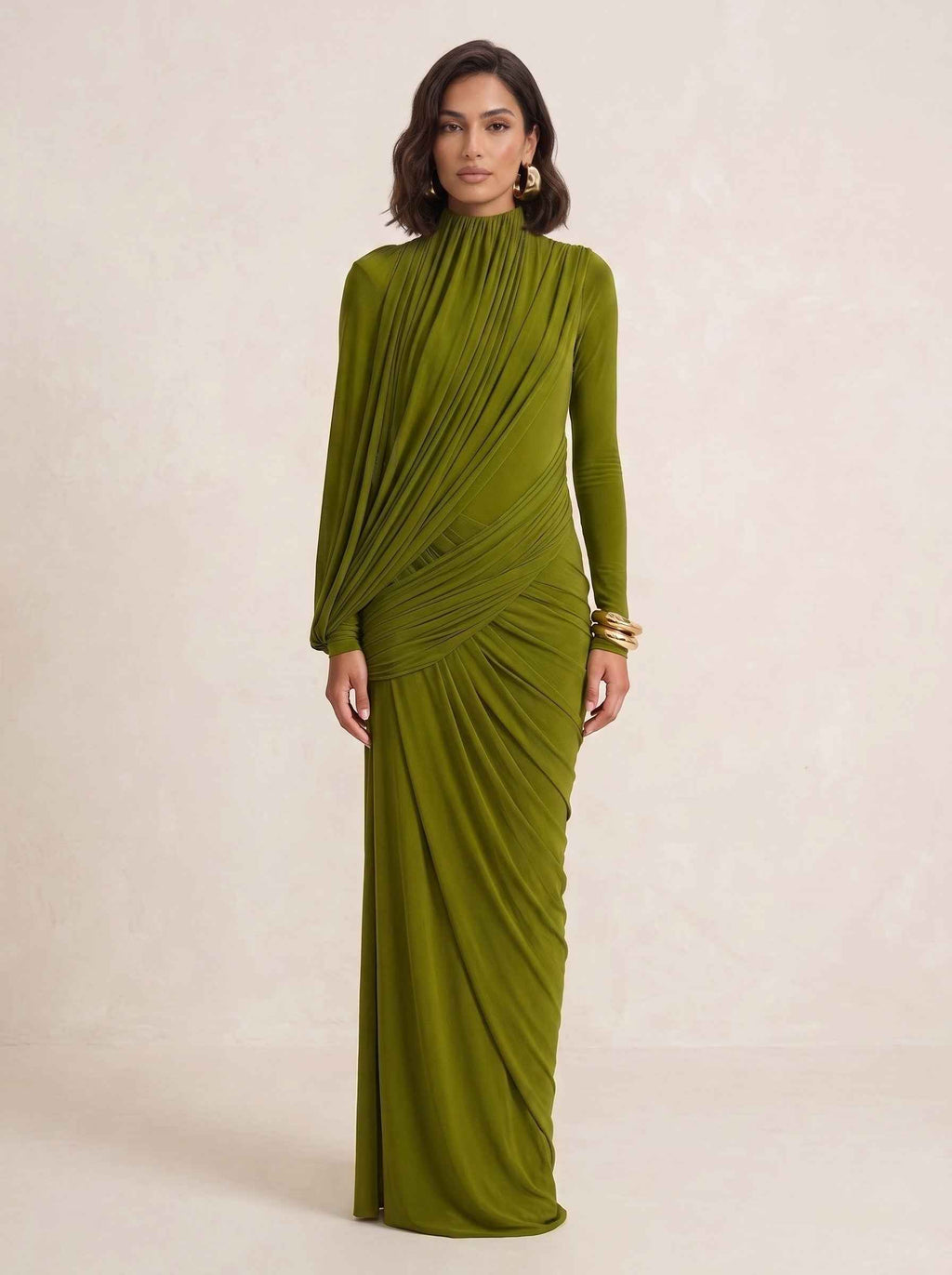 A woman modeling a floor-length olive green gown with a high mock neck, long sleeves, and dramatic draped ruching across the bodice and skirt, posed against a warm beige studio backdrop.
