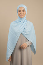 Woman wearing a light blue hijab and gray dress standing in a neutral setting.