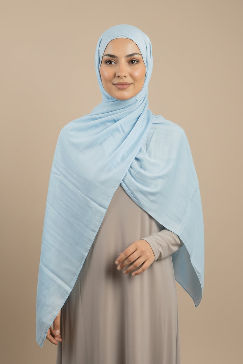 Woman wearing a light blue hijab and gray dress standing in a neutral setting.